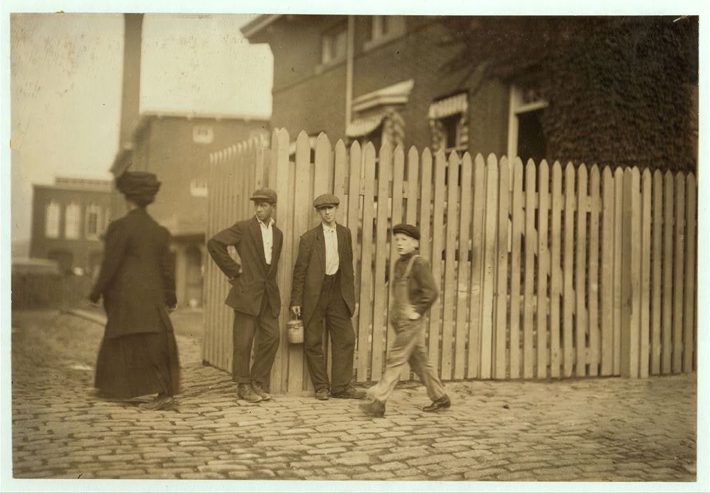 Black and white photograph of four people on a street corner. A teenage boy is walking across.