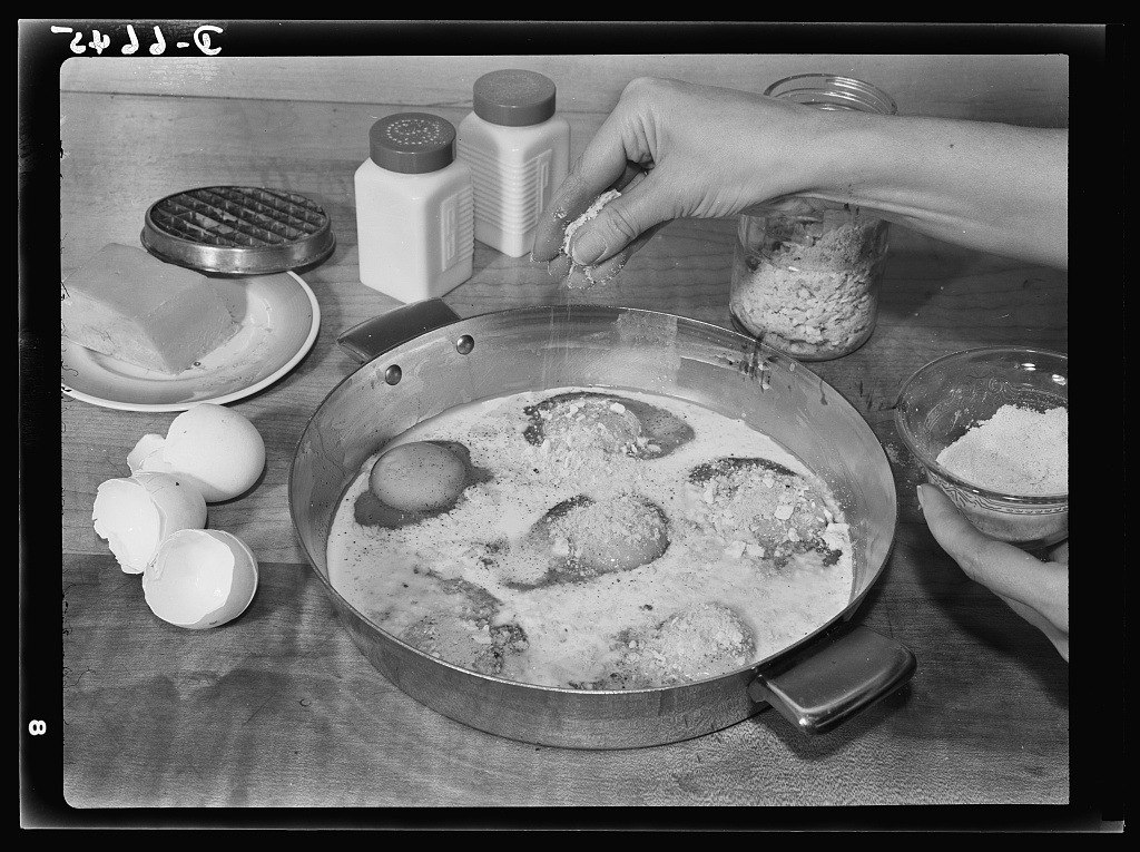 A black and white photograph of a hand sprinkling some sugar (or salt) into a pan with eggs inside of it.