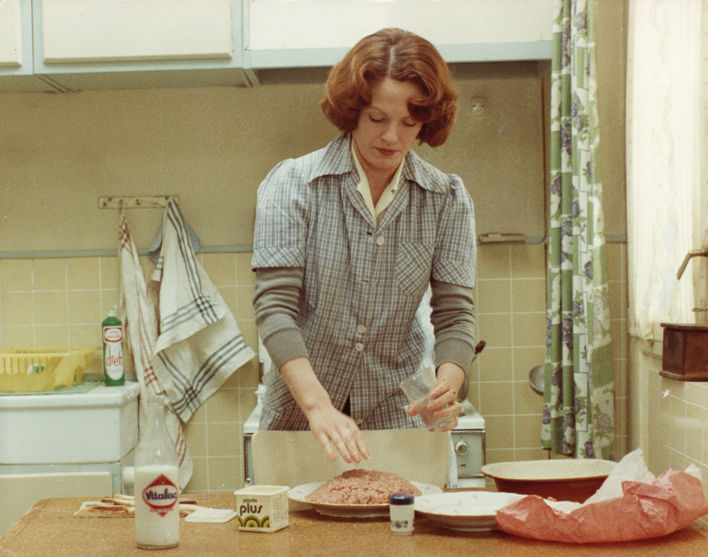 A shot from the movie 'Jeanne Dielman' depicting a woman with wavy brown hair, standing in a kitchen at a table. She is making a meatloaf, various ingredients are on the table: milk, minced beef, salt, an empty plate, and an empty bag.