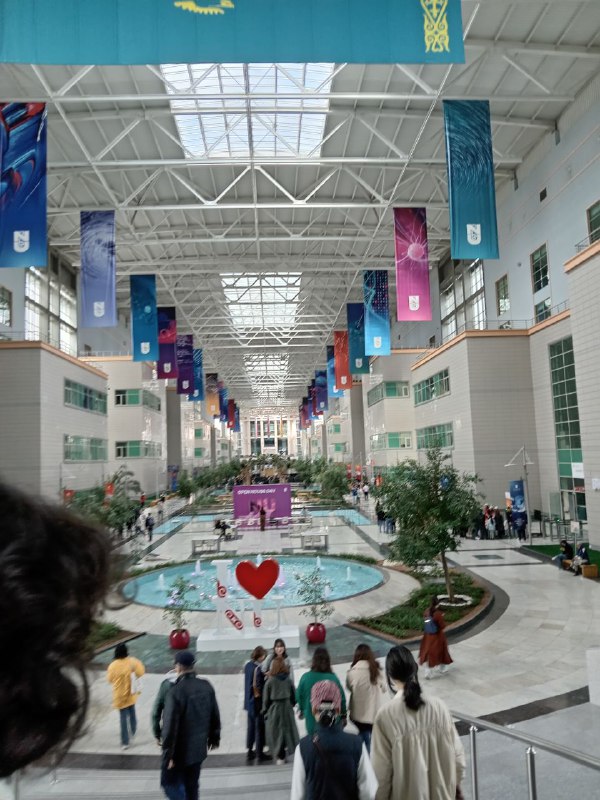 Blurry photograph of a large, open, prolonged main hall. There are coloured-banners hanging from the ceiling, the ceiling has beams and glass. There is a fountain in the centre and a lot of greenery. There are apartment-blocks like buildings on each side.